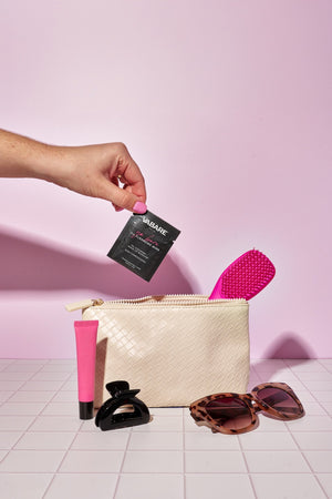 Hand holding a single VABARE Go Bare Oil Cleansing Wipe above a white pouch with cosmetic items on a pink background